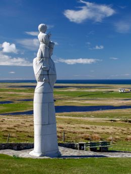The statue called Our lady of the isles, stands proudly tall in the foreground with the flat landscape of South Uist in the background.