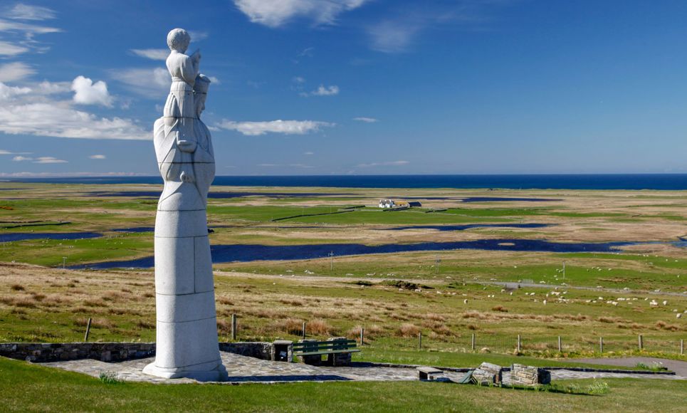 The statue called Our lady of the isles, stands proudly tall in the foreground with the flat landscape of South Uist in the background.