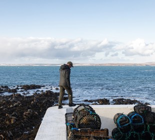 Man walking along a white coastal wall beside a pile of creels in Portnaguran, Isle of Lewis