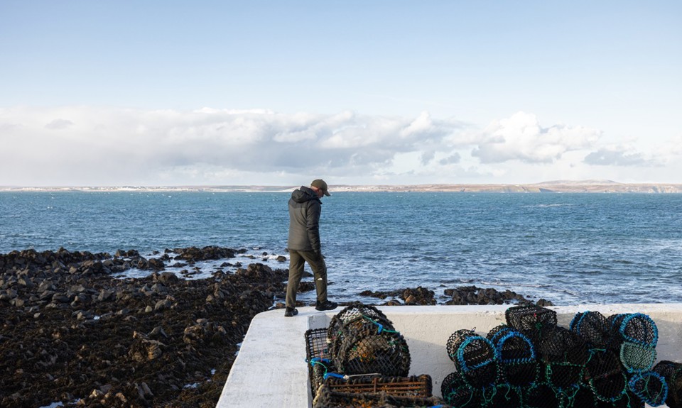 Man walking along a white coastal wall beside a pile of creels in Portnaguran, Isle of Lewis