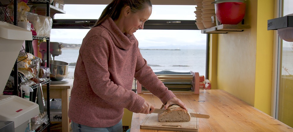 woman in pink sweater slices bread in a yellow kitchen with sea view through window.