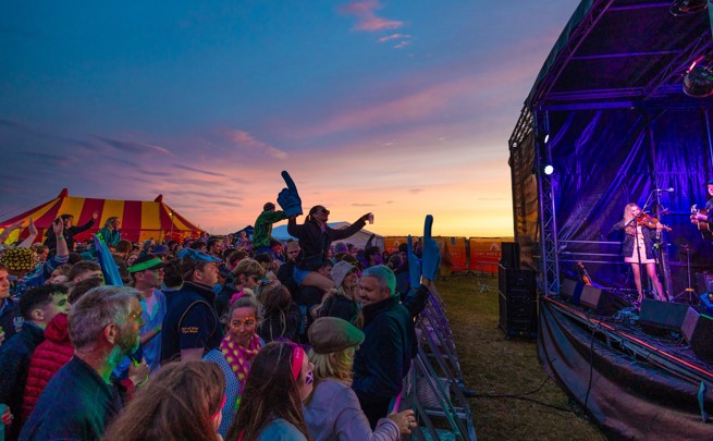 Crowd enjoying an outdoor music performance at sunset, with musicians playing on a stage and a striped festival tent in the background.