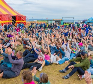 Festivalgoers seated on the grass at Tiree Music Festival, with the main tent and event site visible in the background.