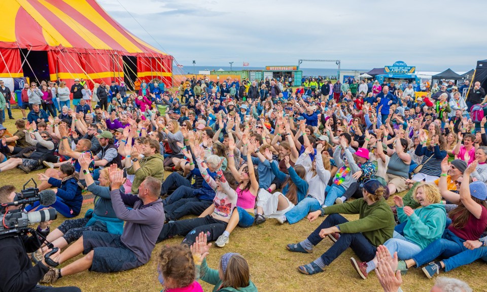 Festivalgoers seated on the grass at Tiree Music Festival, with the main tent and event site visible in the background.