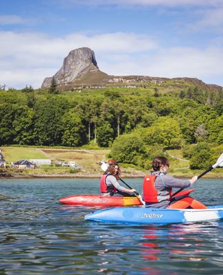 Two people on the water kayaking with trees and the rocky Sgurr of Eigg in the distance.