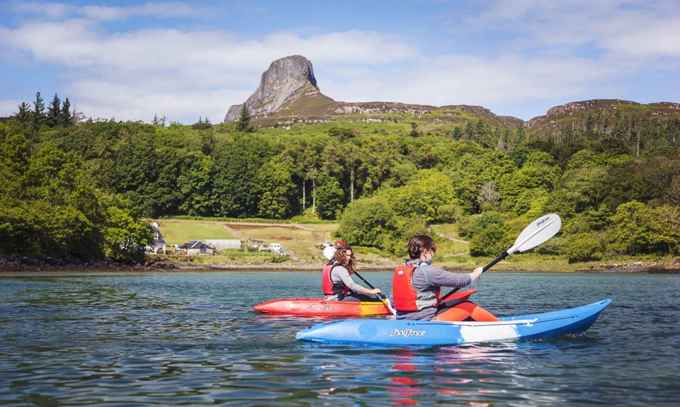 Two people on the water kayaking with trees and the rocky Sgurr of Eigg in the distance.