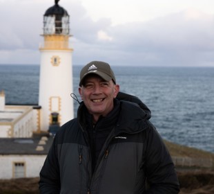 A man smiling standing in front of a lighthouse with the sea behind him. 