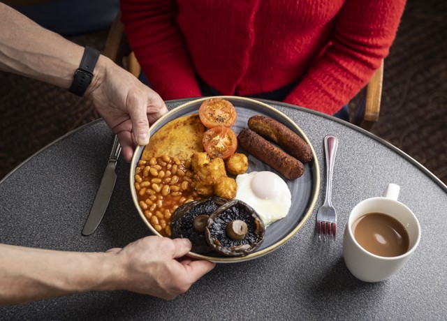 A person in a red sweater is seated at a table, and another person is serving them a plate of food. The plate contains a fried breakfast. 