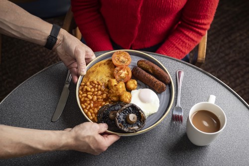 someone placing a vegetarian cooked breakfast in front of a woman wearing a red jumper.