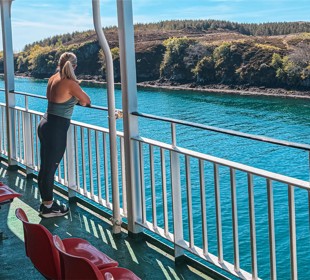 Person stands on ferry deck facing blue water and distant green hills, with red seats lined along the deck railing.