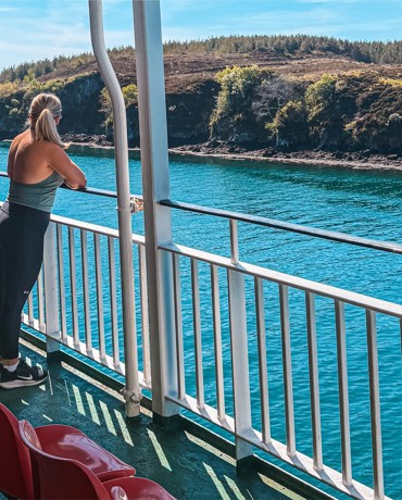 Person stands on ferry deck facing blue water and distant green hills, with red seats lined along the deck railing.