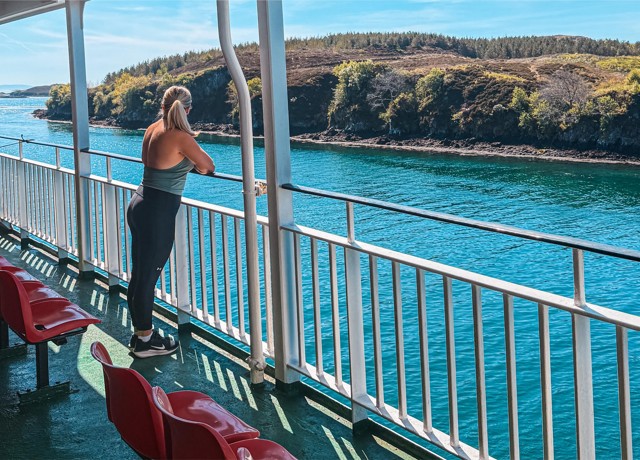 Person stands on ferry deck facing blue water and distant green hills, with red seats lined along the deck railing.