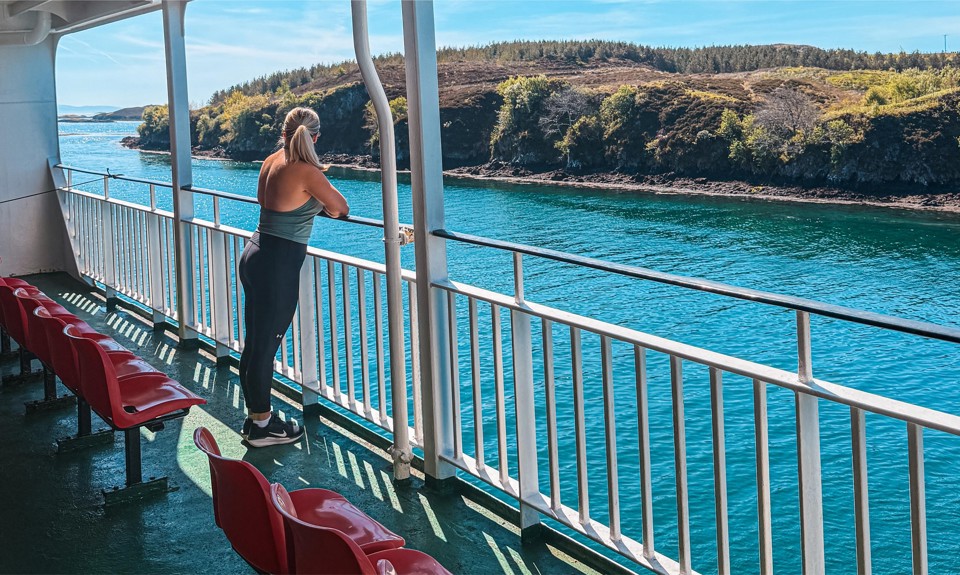 Person stands on ferry deck facing blue water and distant green hills, with red seats lined along the deck railing.