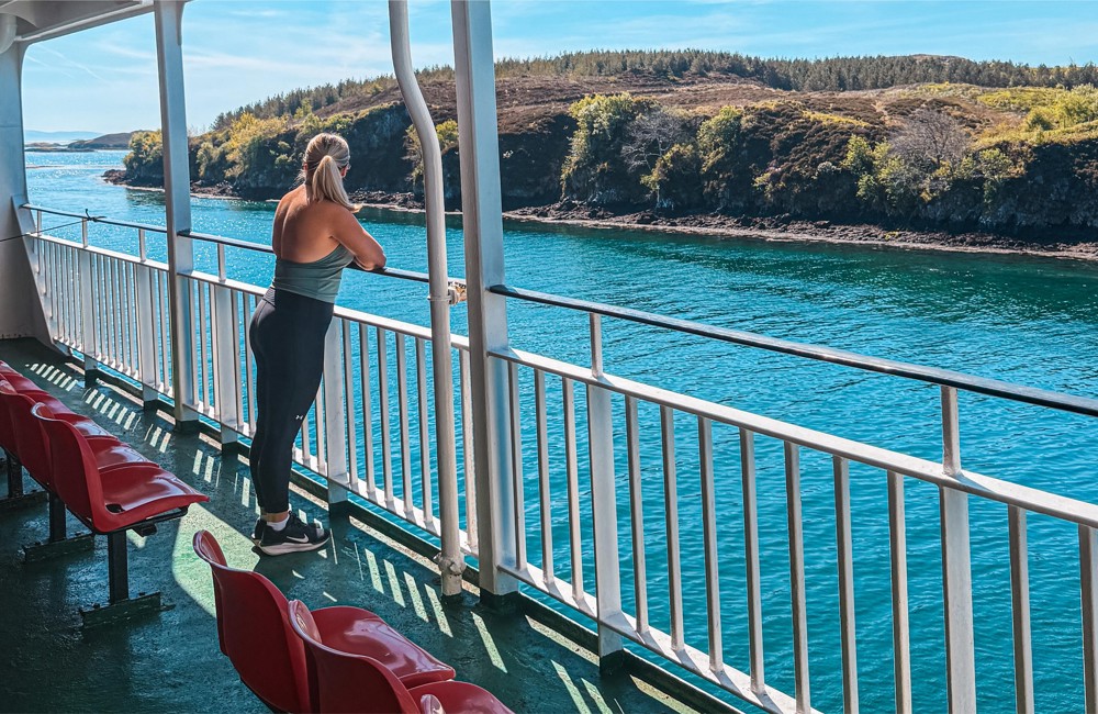 Person stands on ferry deck facing blue water and distant green hills, with red seats lined along the deck railing.