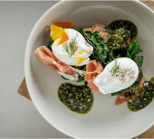 Two poached eggs on greens with prosciutto, garnished with dill and pesto, served on a white plate.