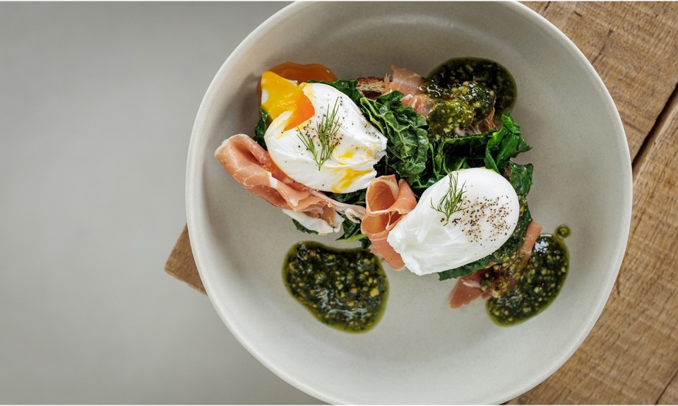 Two poached eggs on greens with prosciutto, garnished with dill and pesto, served on a white plate.