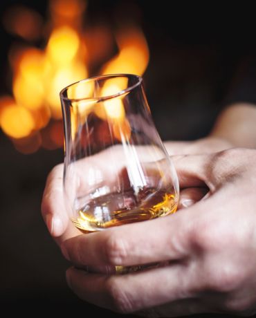 A person with a glass of whisky held in two hands with a flames from a indoor fire in the background