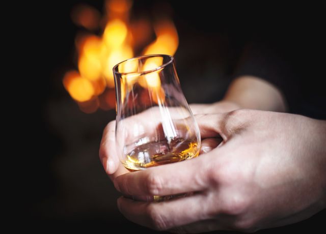 A person with a glass of whisky held in two hands with a flames from a indoor fire in the background