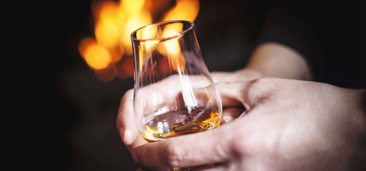 A person with a glass of whisky held in two hands with a flames from a indoor fire in the background