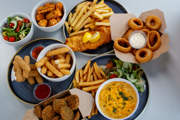 Assorted side dishes including fish and chips, onion rings, mozzarella sticks, breaded spinach, grilled corn, mac and cheese, and fresh salad.