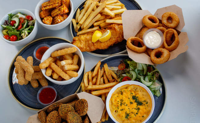 Assorted side dishes including fish and chips, onion rings, mozzarella sticks, breaded spinach, grilled corn, mac and cheese, and fresh salad.