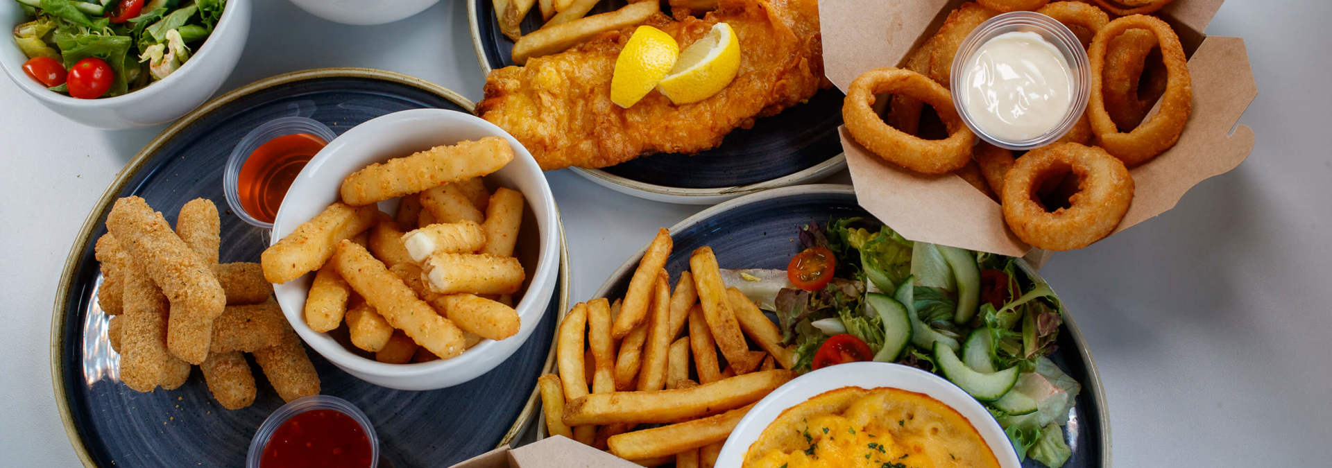 Assorted side dishes including fish and chips, onion rings, mozzarella sticks, breaded spinach, grilled corn, mac and cheese, and fresh salad.