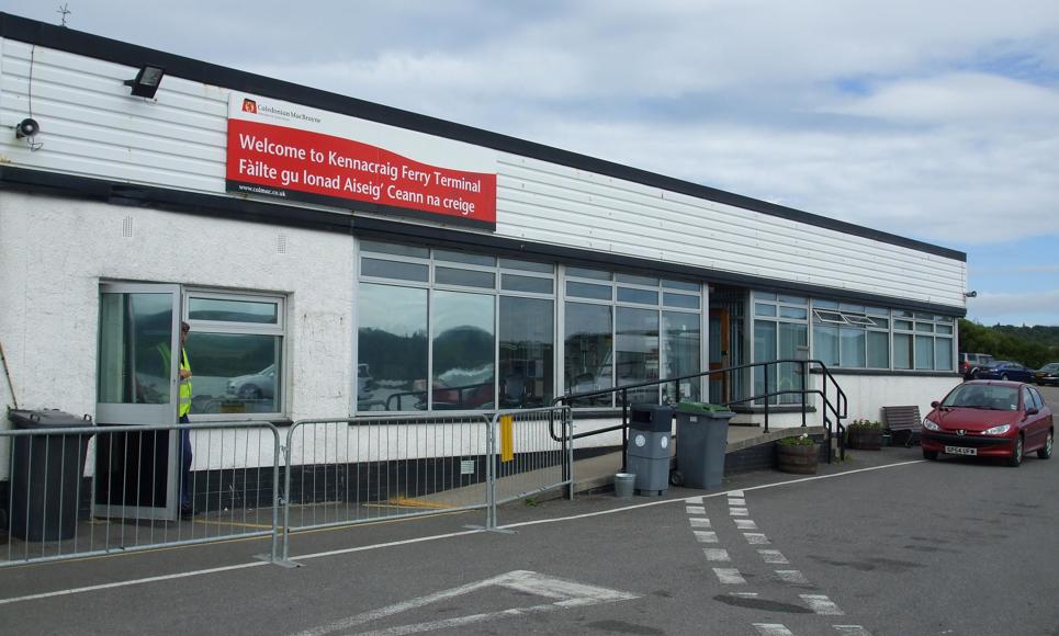 Kennacraig Ferry Terminal with a red bilingual welcome sign in English and Gaelic, accessible parking area, and a red car nearby.