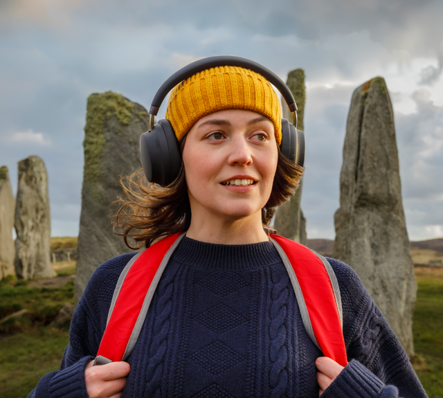 Brunette female with yellow hat, red backpack and black over ear headphones standing in front of ancient standing stones