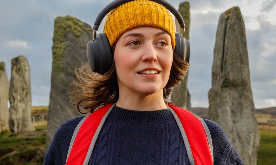 Brunette female with yellow hat, red backpack and black over ear headphones standing in front of ancient standing stones