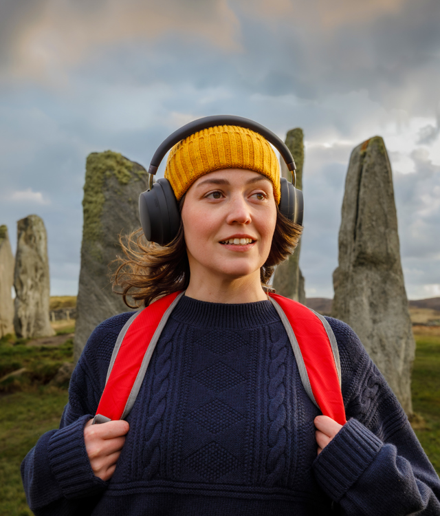 Brunette female with yellow hat, red backpack and black over ear headphones standing in front of ancient standing stones