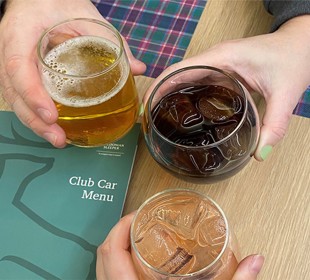 Three hands holding beer, cola, and a cocktail over a table with two green menus which say 'Club Car Menu' on the front.