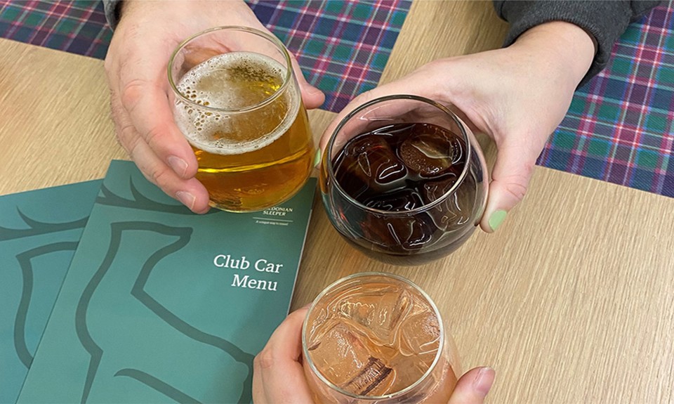 Three hands holding beer, cola, and a cocktail over a table with two green menus which say 'Club Car Menu' on the front.