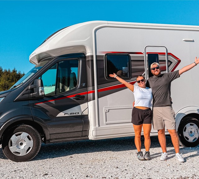 Two people with arms raised stand in front of a white and black motorhome parked on gravel, with trees and mountains in the background.
