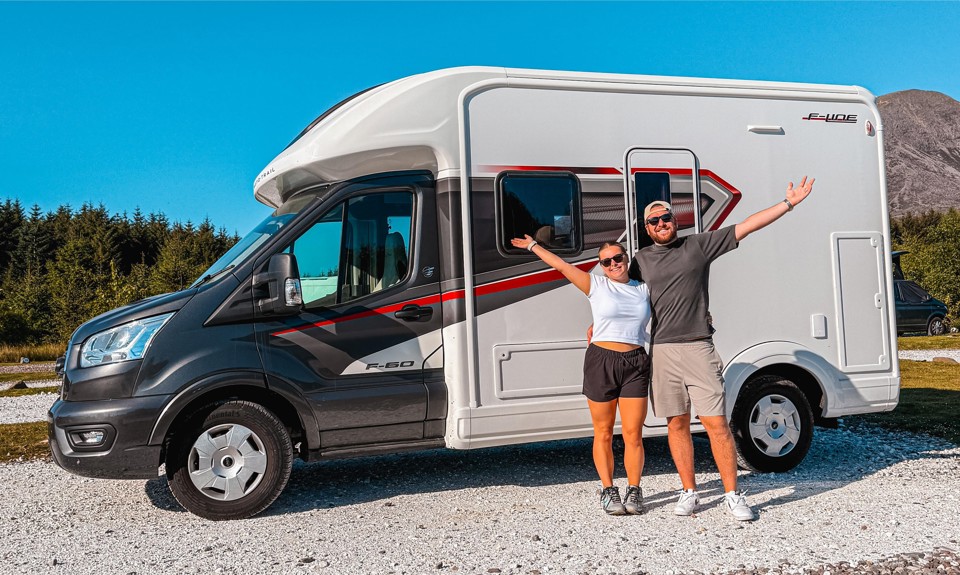 Two people with arms raised stand in front of a white and black motorhome parked on gravel, with trees and mountains in the background.
