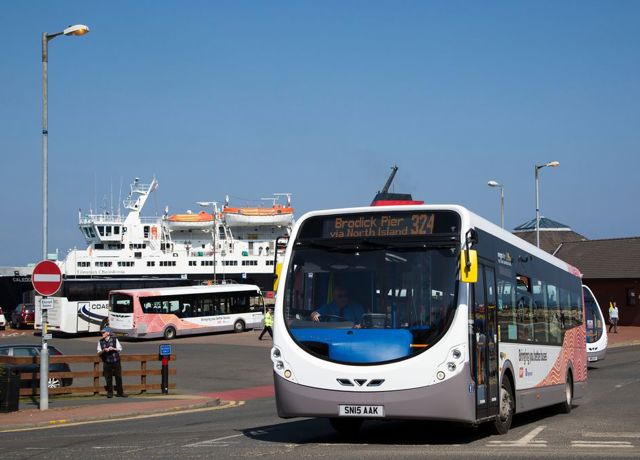 Bus 324 to Brodick Pier driving near a ferry terminal, with a large ferry and another bus visible in the background.