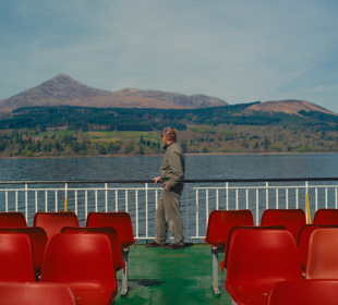 Passenger standing on a ferry deck with red seats, overlooking calm water and mountains across the shoreline.