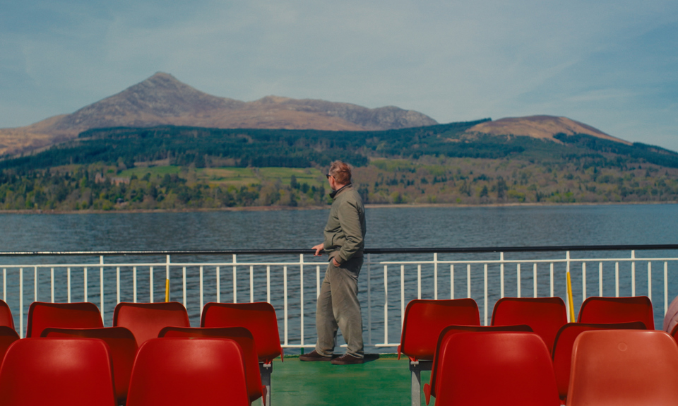 Passenger standing on a ferry deck with red seats, overlooking calm water and mountains across the shoreline.