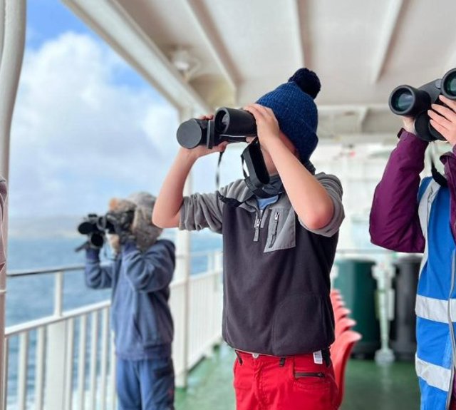 Group of kids outside on a ferry looking out to sea with binoculars 