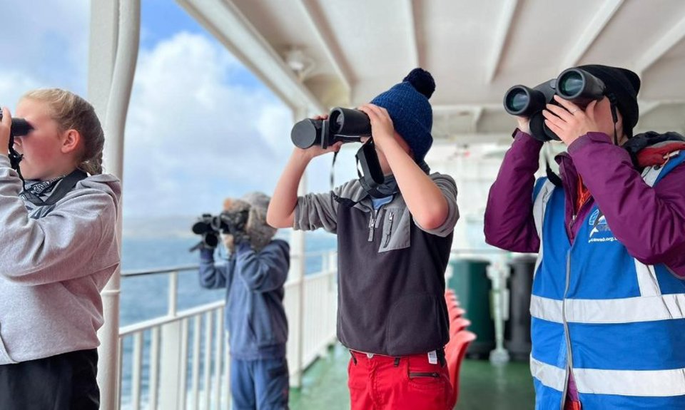 Group of kids outside on a ferry looking out to sea with binoculars 