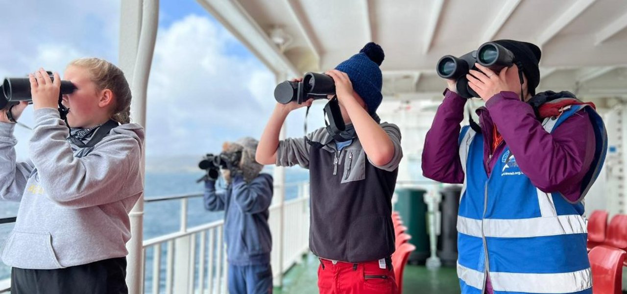 Group of kids outside on a ferry looking out to sea with binoculars 