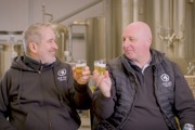 Two men in brewery uniforms clink beer glasses in front of brewing equipment.