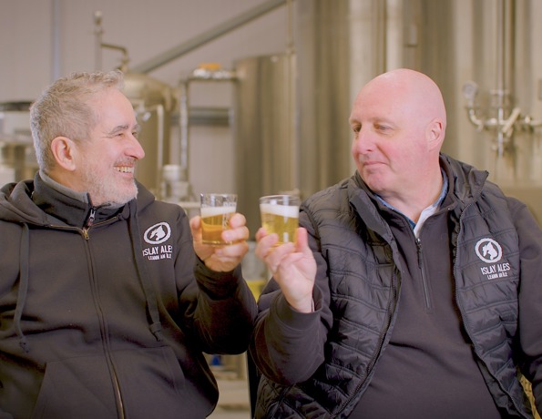 Two men in brewery uniforms clink beer glasses in front of brewing equipment.