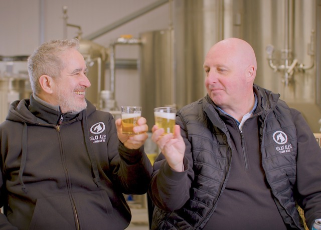 Two men in brewery uniforms clink beer glasses in front of brewing equipment.