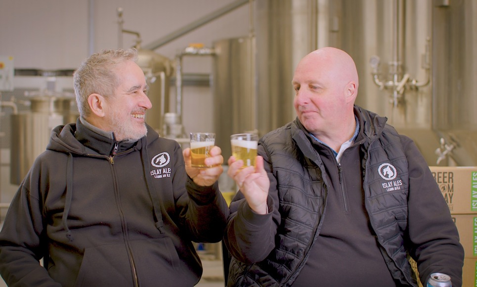Two men in brewery uniforms clink beer glasses in front of brewing equipment.