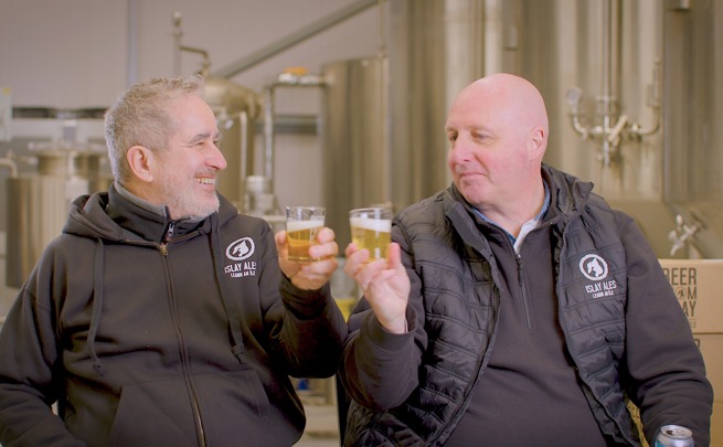 Two men in brewery uniforms clink beer glasses in front of brewing equipment.