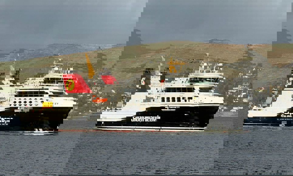 Caledonian MacBrayne ferry ‘Isle of Islay’ sailing across a calm sea with hills and houses in the background.