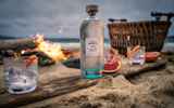 A selction of products from Isle of Harris Distillers set up on a beach next to a fire. Products include Harris Gin, and three glasses with a garnish.