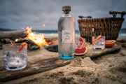 A selction of products from Isle of Harris Distillers set up on a beach next to a fire. Products include Harris Gin, and three glasses with a garnish.