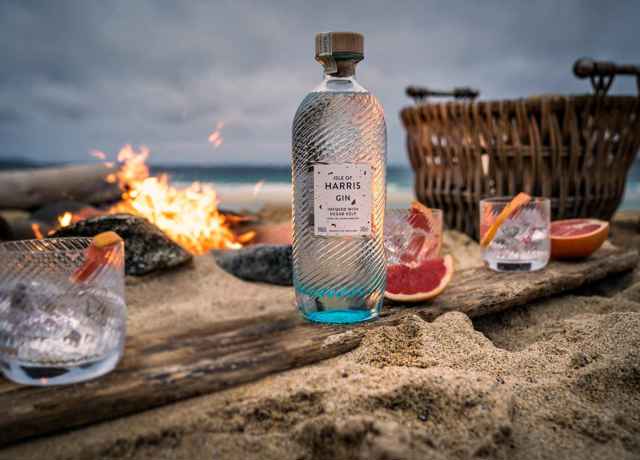 A selction of products from Isle of Harris Distillers set up on a beach next to a fire. Products include Harris Gin, and three glasses with a garnish.