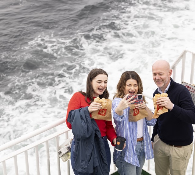 two ladies and a man taking a selfie out on deck with the sea behind them